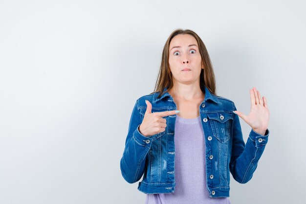 Jonge dame wijst naar haar opgeheven hand in t-shirt, jas en kijkt verbaasd. vooraanzicht.