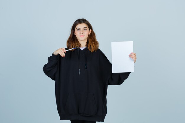 Jonge dame wijst papier met pen in oversized hoodie, broek en ziet er zelfverzekerd uit, vooraanzicht.