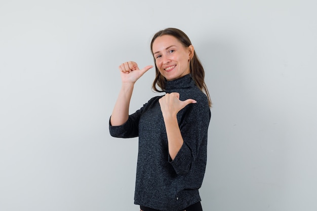 Jonge dame wijst terug met duimen in casual overhemd en ziet er vrolijk uit. vooraanzicht.