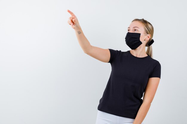 Jonge dame wijst weg in t-shirt, broek, medisch masker en ziet er zelfverzekerd uit