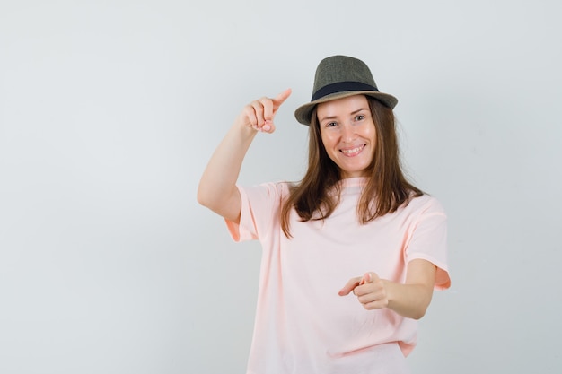 Jonge dame wijzend in roze t-shirt, hoed en op zoek gelukkig, vooraanzicht.