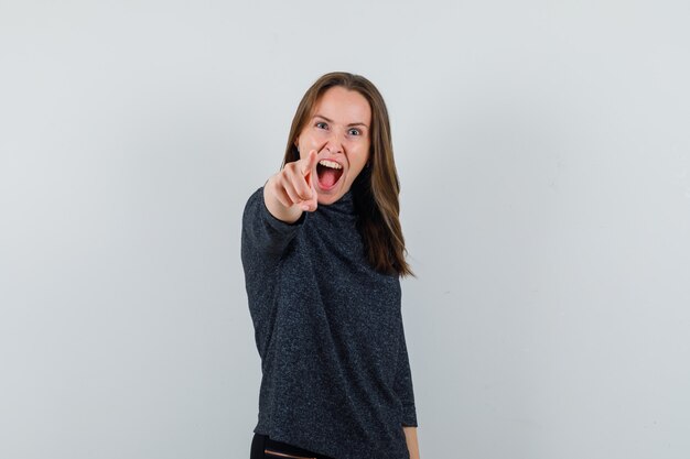 Jonge dame wijzend op camera in casual shirt en op zoek gek. vooraanzicht.