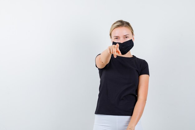 Jonge dame wijzend op camera in zwart t-shirt, masker en op zoek zelfverzekerd. vooraanzicht.