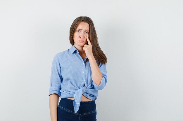 Jonge dame wijzend op onderste ooglid in blauw shirt, broek en op zoek depressief