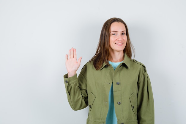 Jonge dame zwaait met de hand voor begroeting in t-shirt, jas en ziet er schattig uit. vooraanzicht.