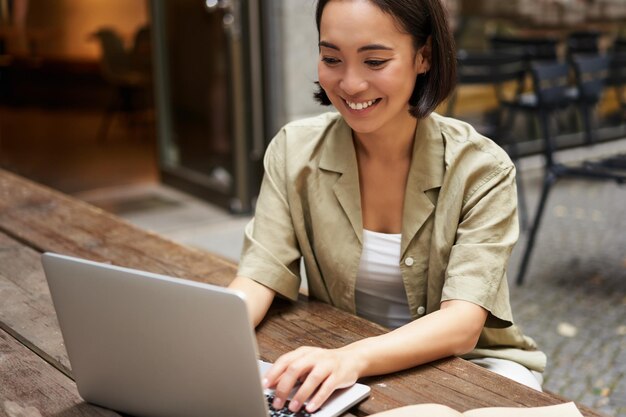 Jonge digitale nomadenvrouw die op afstand werkt vanuit café typend op laptop buiten zitten en glimlachen