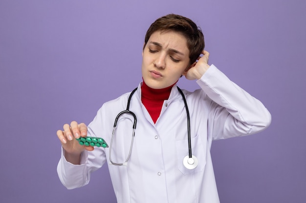 Jonge dokter in witte jas met stethoscoop om nek met blaar met pillen die ernaar kijkt verward met hand op haar hoofd die op paars staat