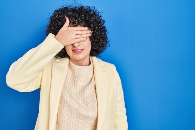 Jonge donkerbruine vrouw met krullend haar die zich over blauwe achtergrond bevindt die ogen met hand behandelt, ernstig en droevig kijkend. zichtloos, verbergen en afwijzen concept