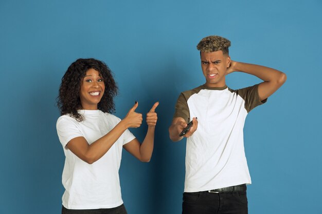 Jonge emotionele Afro-Amerikaanse man en vrouw die zich voordeed op blauwe achtergrond. Mooi paar. Concept van menselijke emoties, gezichtsuitdrukkingen, relaties, advertentie. Kijk samen tv, haar favoriete kanaal.