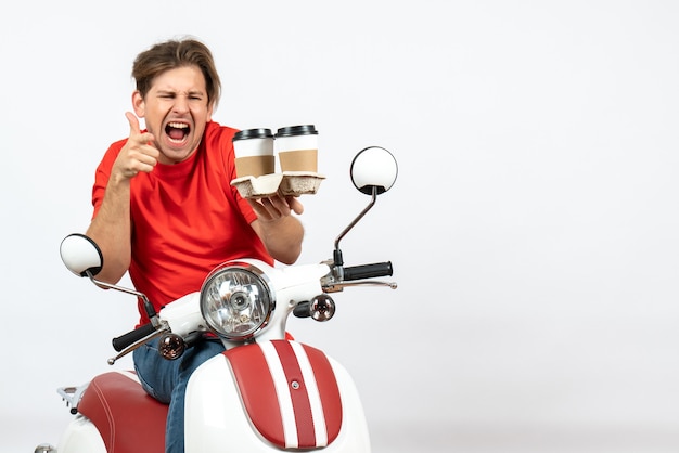 Jonge emotionele ambitieuze koerierskerel in rood uniform zittend op scooter met bestellingen en ok gebaar maken op gele muur