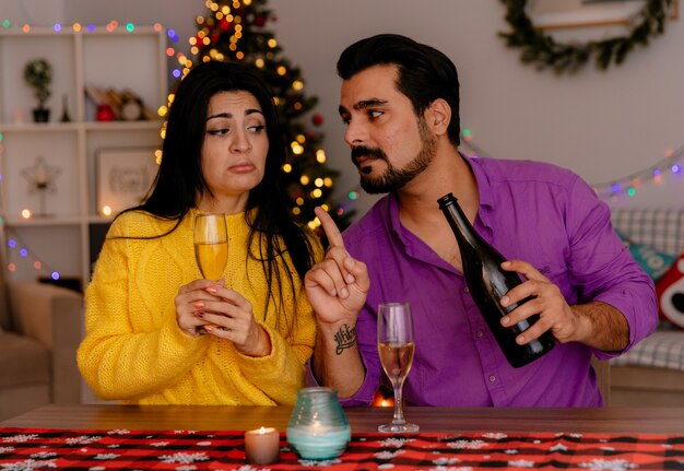Jonge en mooie paar man en vrouw zitten aan de tafel met glazen champagne vieren kerst samen in kerst ingerichte kamer met kerstboom in de muur