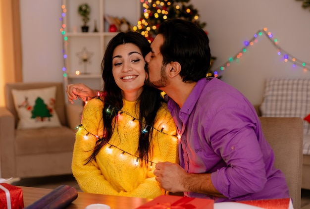 Jonge en mooie paar man en vrouw zitten aan de tafel met slinger man zoenen zijn vriendin in kerst ingerichte kamer met kerstboom in de muur