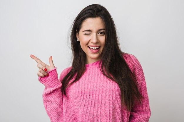 Jonge en mooie vrouw in roze warme trui, natuurlijke uitstraling, glimlachen, wijzende vinger opzij, knipogen, portret aan, geïsoleerd, lang haar, grappige gezichtsuitdrukking, positieve stemming