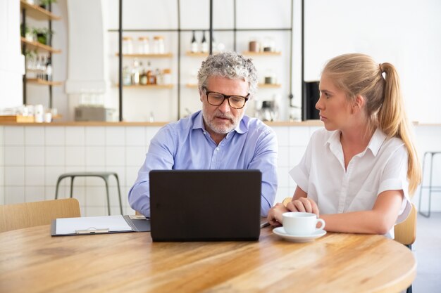 Jonge en volwassen collega's bijeen tijdens co-working, zittend op opengeklapte laptop, inhoud bespreken