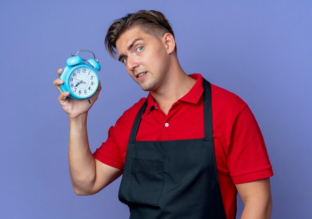 Jonge ernstige blonde mannelijke kapper in uniform houdt wekker geïsoleerd op violette ruimte met kopie ruimte