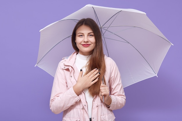 Jonge Europese vrouw die een paraplu houdt die over lila muur wordt geïsoleerd, hand op borst houdt glimlacht oprecht, dankbaar is, haar prettige houding uitdrukt.