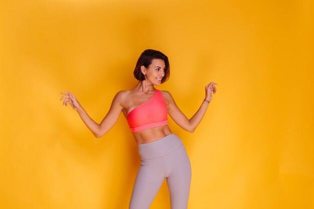 Jonge fit vrij sterke vrouw gekleed in sportkleding, stijlvolle top en legging, poseert tegen gele muur