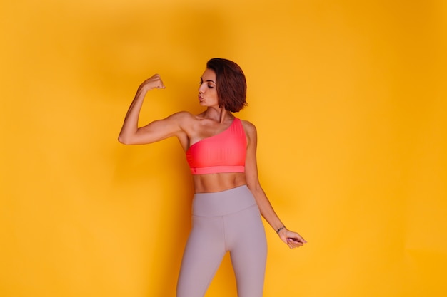Jonge fit vrij sterke vrouw gekleed in sportkleding, stijlvolle top en legging, poseert tegen gele muur