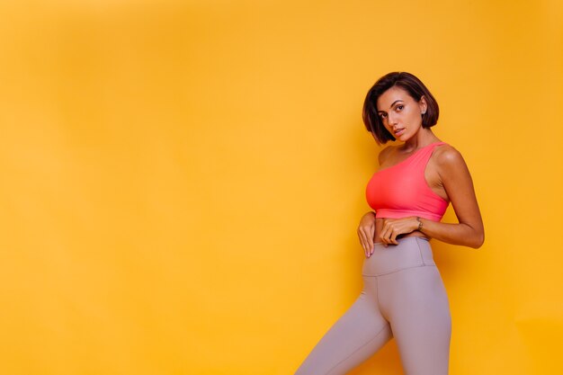 Jonge fit vrij sterke vrouw gekleed in sportkleding, stijlvolle top en legging, poseert tegen gele muur