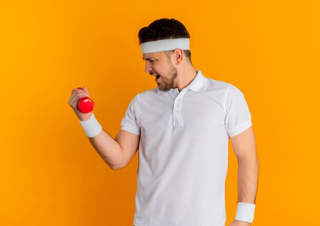 Jonge fitness man in wit overhemd met hoofdband doen oefeningen met halter op zoek gespannen staande over oranje achtergrond