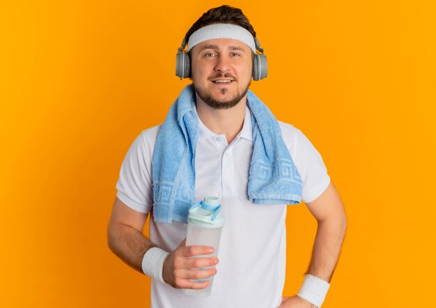 Jonge fitness man in wit overhemd met hoofdband en handdoek om de nek met fles water camera kijken met zelfverzekerde uitdrukking staande over oranje achtergrond
