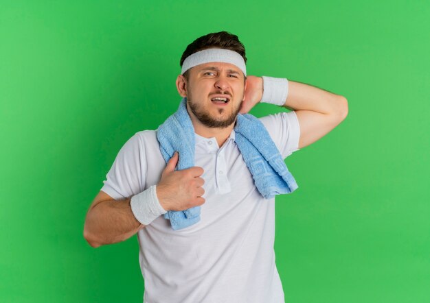 Jonge fitness man in wit overhemd met hoofdband en handdoek om zijn nek camera kijken moe en uitgeput na training staande op groene achtergrond