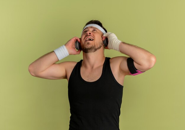 Jonge fitness man met hoofdband en koptelefoon luisteren naar muziek staande over olijf achtergrond
