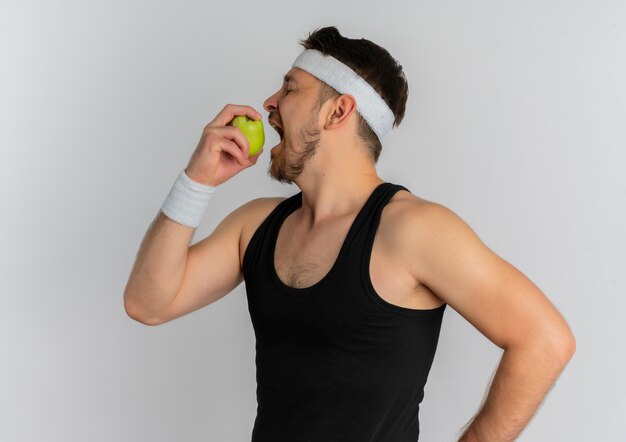 Jonge fitness man met hoofdband met groene appel bijten staande op witte achtergrond