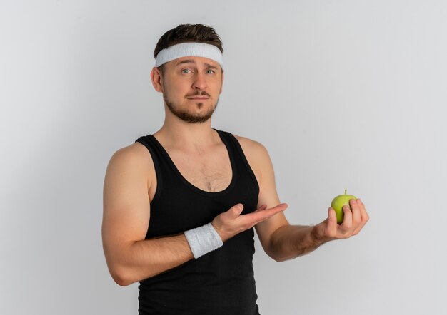 Jonge fitness man met hoofdband met groene appels presenteren het met arm oh hand kijken met ernstig gezicht staande op witte achtergrond