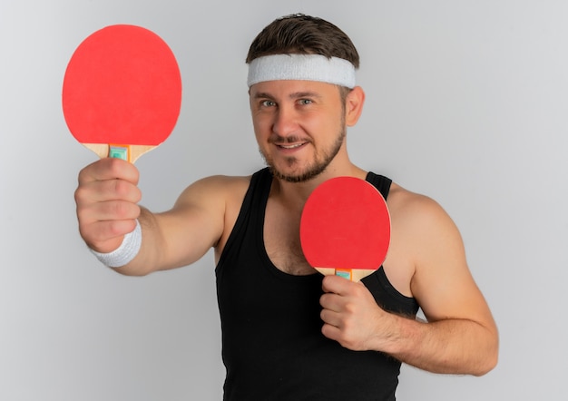 Jonge fitness man met hoofdband met twee rackets camera kijken met glimlach op gezicht staande op witte achtergrond