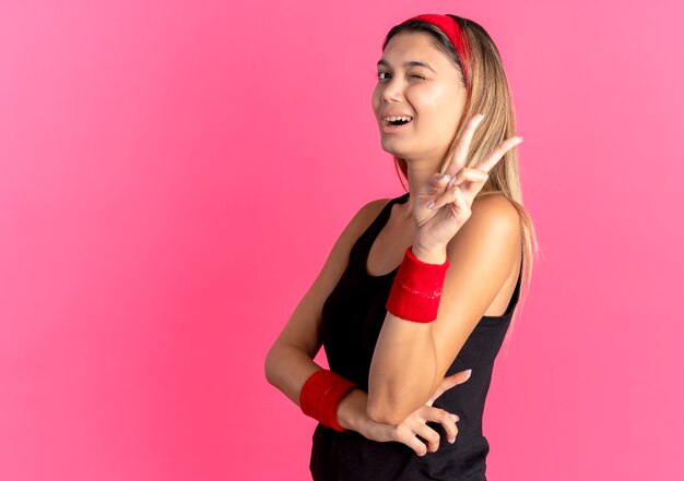 Jonge fitness meisje in zwarte sportkleding en rode hoofdband glimlachend en knipogen weergegeven: overwinningsteken over roze