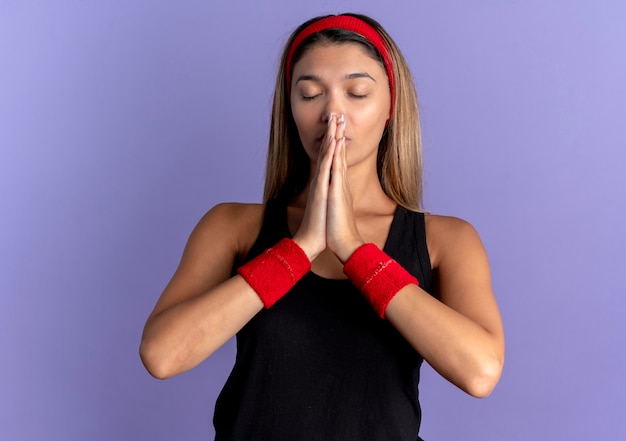 Jonge fitness meisje in zwarte sportkleding en rode hoofdband palmen bij elkaar te houden als bidden met hoop expressie met gesloten ogen over blauw