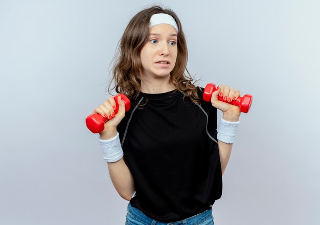 Jonge fitness meisje in zwarte sportkleding met hoofdband uit te werken met halters op zoek verward staande over witte muur