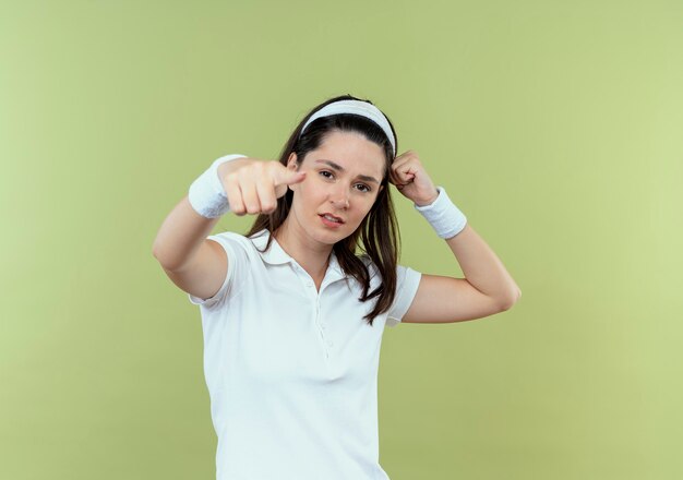 Jonge fitness vrouw die in hoofdband zelfverzekerd kijkt wijzend met vinger die zich over lichte muur bevindt