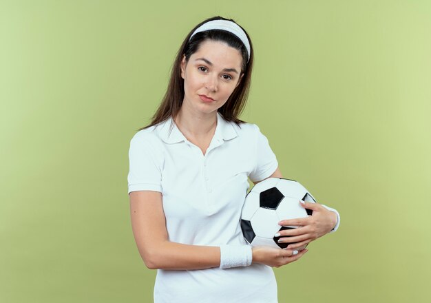 Jonge fitness vrouw in het voetbal van de hoofdbandholding die camera met zekere uitdrukking bekijkt die zich over lichte achtergrond bevindt
