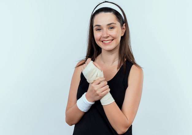 Jonge fitness vrouw in hoofdband aanraken haar verbonden pols camera kijken met glimlach op gezicht staande op witte achtergrond