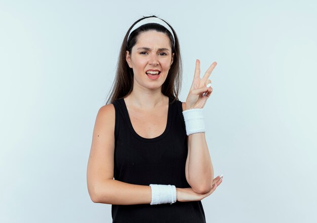 Jonge fitness vrouw in hoofdband kijken camera glimlachend weergegeven: overwinning teken staande op witte achtergrond