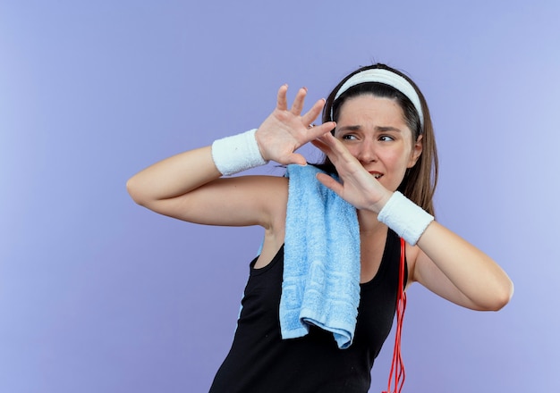 Gratis foto jonge fitness vrouw in hoofdband met handdoek op haar schouder bang verdediging gebaar met handen permanent over blauwe achtergrond