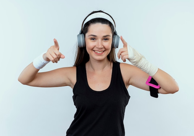 jonge fitness vrouw in hoofdband met koptelefoon en smartphone armband lachend met blij gezicht wijzend met wijsvingers naar de zijkant staande over witte muur