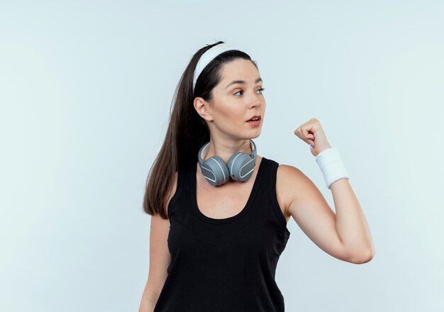 Jonge fitness vrouw in hoofdband met koptelefoon opzij kijken met gebalde vuist staande op witte achtergrond