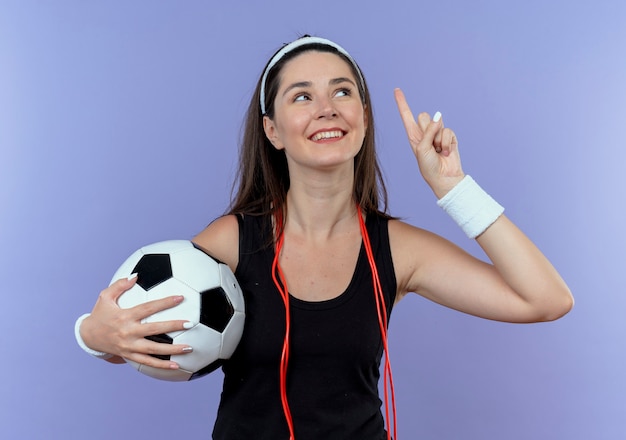 Jonge fitness vrouw in hoofdband met springtouw om nek met voetbal omhoog met vinger opzoeken glimlachend met nieuw idee staande over blauwe muur