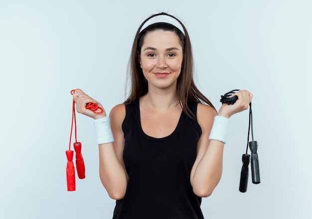 Jonge fitness vrouw in hoofdband met springtouwen kijken camera glimlachend staande op witte achtergrond