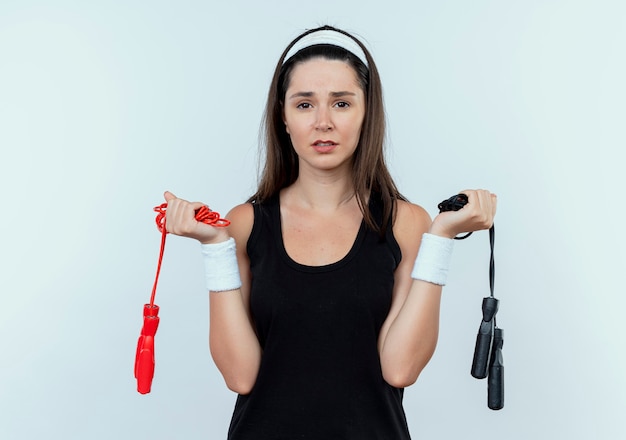 Jonge fitness vrouw in hoofdband met springtouwen kijken camera met droevige uitdrukking op gezicht staande op witte achtergrond