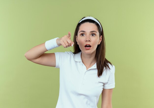 Jonge fitness vrouw in hoofdband op zoek verrast wijzend met vinger op iets staande over lichte achtergrond