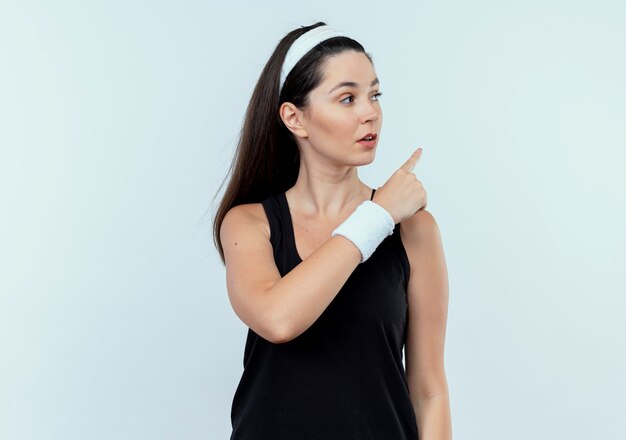 Jonge fitness vrouw in hoofdband opzij kijken met ernstig gezicht wijzend met ndex vinger naar iets staande op een witte achtergrond