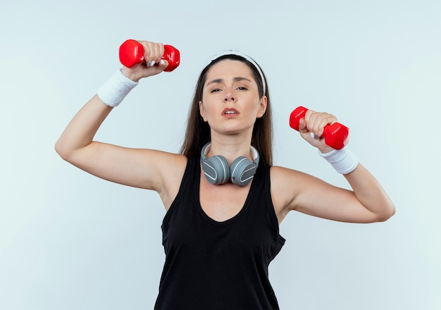 Jonge fitness vrouw in hoofdband uit te werken met halters op zoek moe staande op witte achtergrond