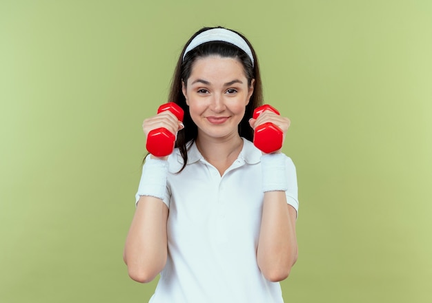 Jonge fitness vrouw in hoofdband uit te werken met halters op zoek zelfverzekerd glimlachend staande over lichte muur