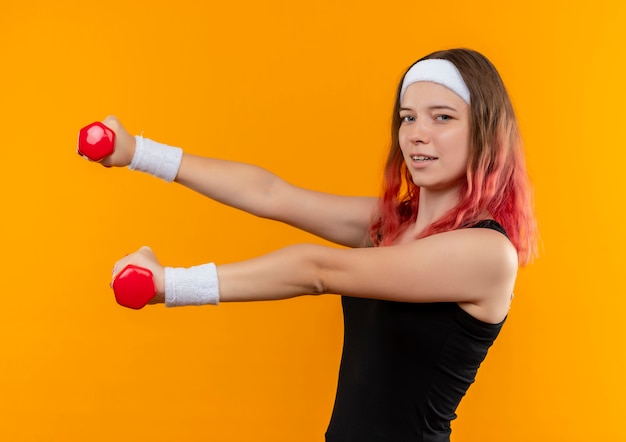 Jonge fitness vrouw in sportkleding met behulp van halters doen oefeningen op zoek zelfverzekerd glimlachend staande over oranje muur
