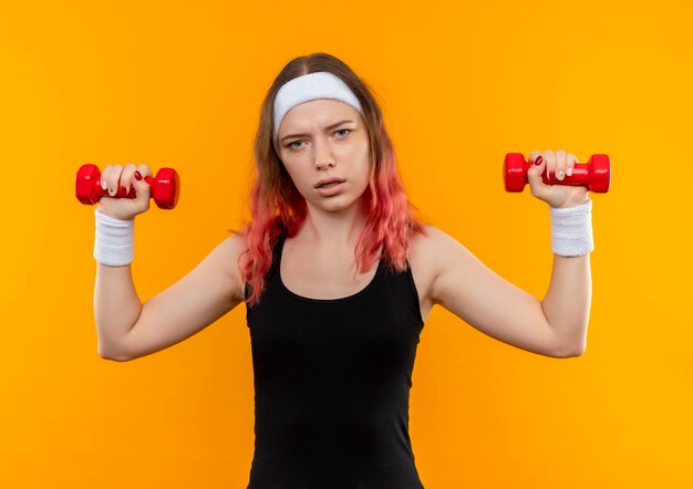 Jonge fitness vrouw in sportkleding met behulp van halters doen oefeningen op zoek zelfverzekerd staande over oranje muur