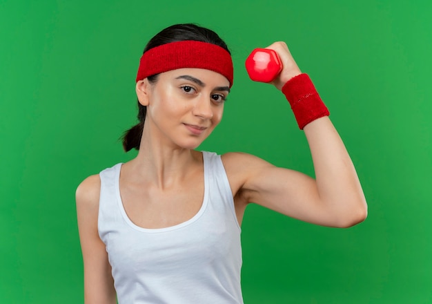 Jonge fitness vrouw in sportkleding met de halter die van de hoofdbandholding hand het glimlachen zelfverzekerd over groene muur opheffen